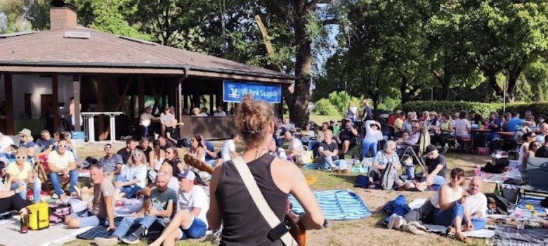 Eine Person mit Zopf und schwarzer Kleidung spielt Gitarre vor einer großen Gruppe von Menschen, die auf einer Wiese bei Sonnenschein sitzen und einem Open-Air-Konzert zuhören.