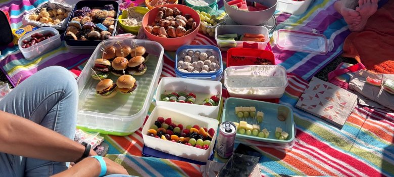 Auf einer bunten Decke im Freien sind verschiedene Snacks, Gemüse, Getränke und Süßigkeiten für ein Picknick angerichtet