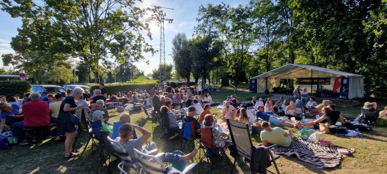 Viele Menschen sitzen bei sonnigem Wetter auf Stühlen und Decken auf einer Wiese vor einem Pavillon und genießen eine Veranstaltung im Freien.
