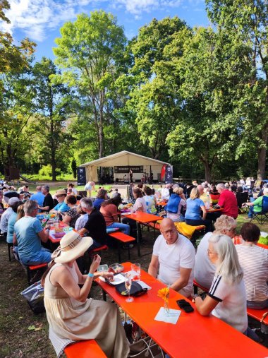 Viele Menschen sitzen an Biertischgarnituren im Freien vor einer Bühne an der Grillhütte mit großen Bäumen und blauem Himmel.  